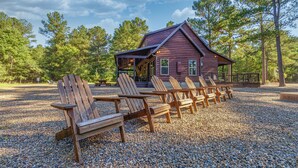 A cabin with a porch, surrounded by wooden Adirondack chairs on a gravel area.