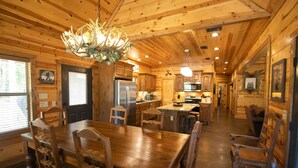 A wooden cabin interior with a dining table, chairs, a chandelier, kitchen area, and a living space with a couch.