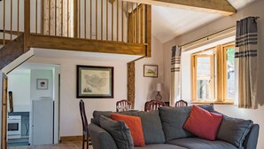 A cozy living room with a grey sofa, wooden beams, a dining area, and a kitchen.