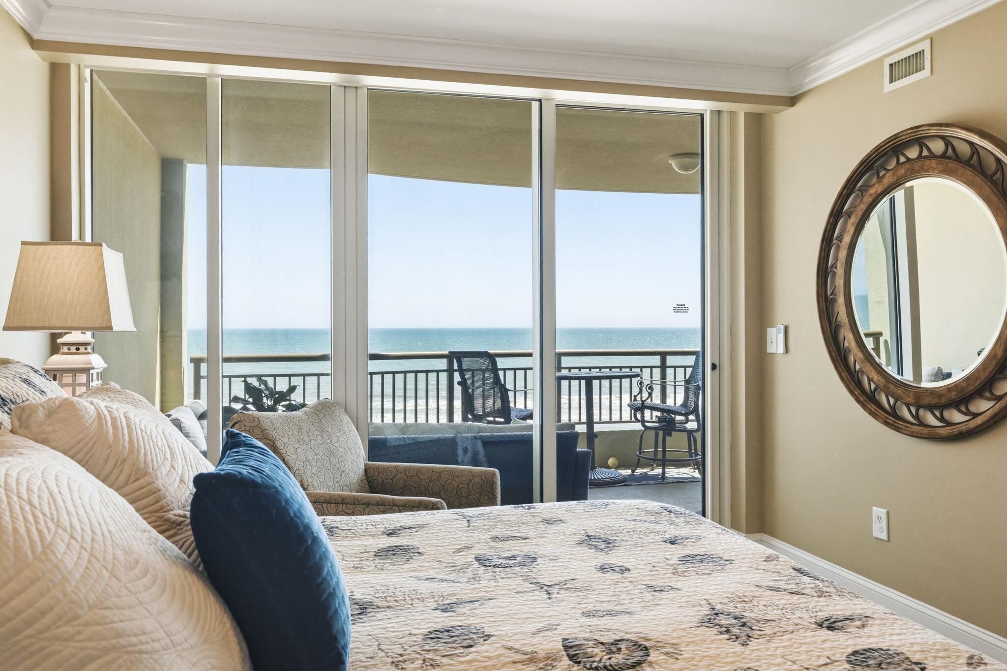 A hotel room with a bed, a large window, a balcony, and a round mirror on the wall.