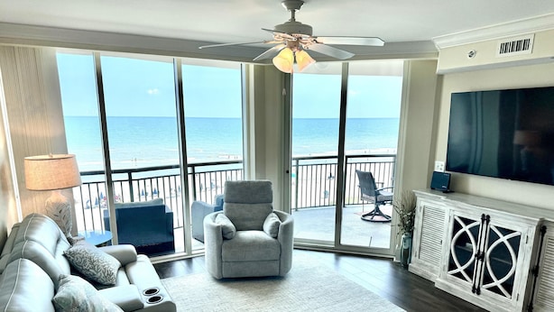 A spacious living room with a large sofa, a chair, and a television. It features a balcony with a view of the ocean. - MVG 509 Oceanfront Club floor (North Myrtle Beach)