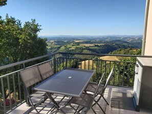 Ein Balkon mit Tisch und Stühlen, der Blick auf ein Tal mit Feldern und Bäumen bietet.