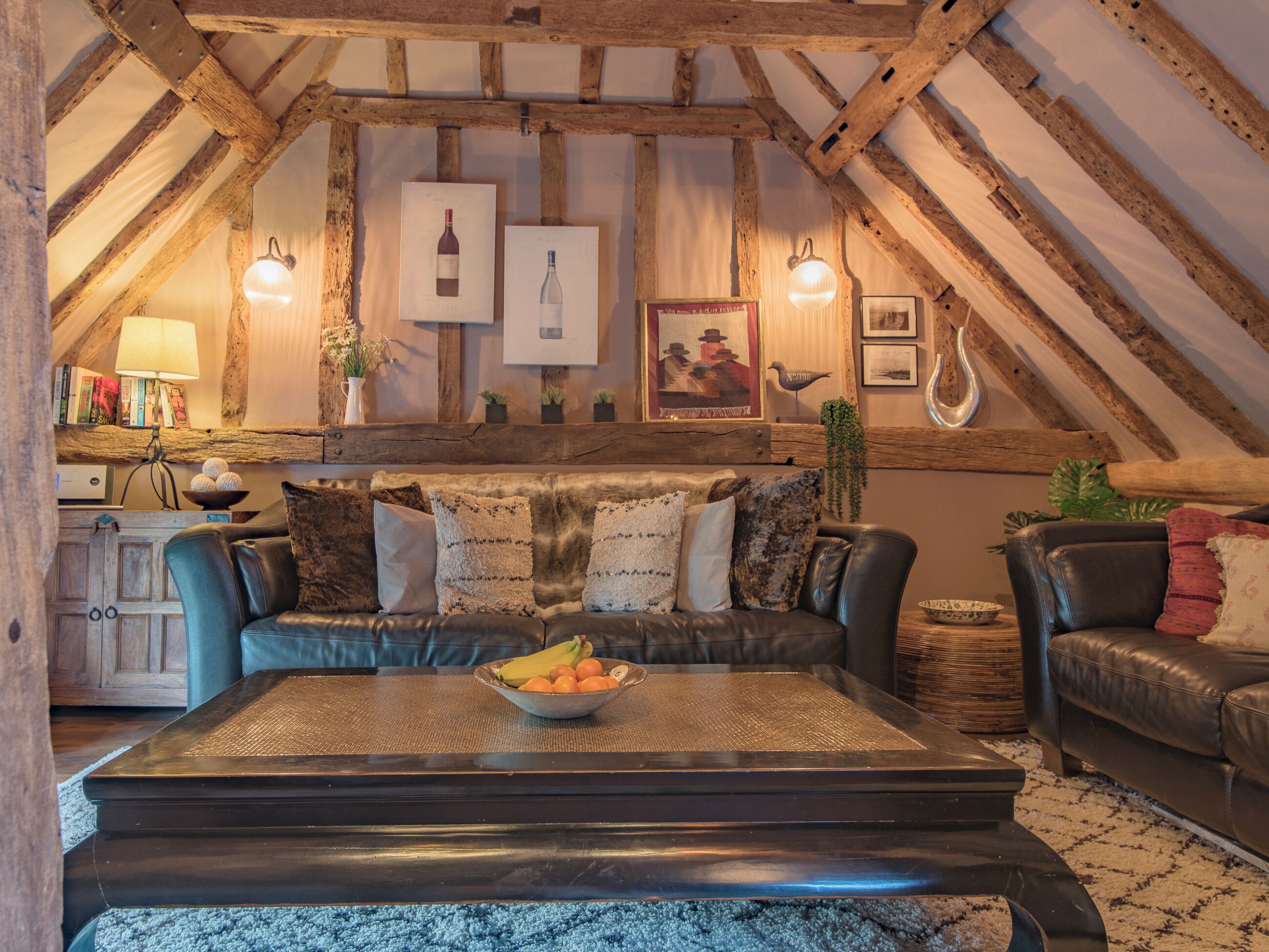 A cozy loft-style living room with exposed wooden beams, a leather sofa, and a coffee table with fruit.
