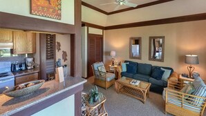 A living room with a sofa, coffee table, and wicker chairs. There is a kitchen area with a stove and cabinets. - Poipu Ground Floor Ease w/Kitchen – Kiahuna 2089 (KOLOA)