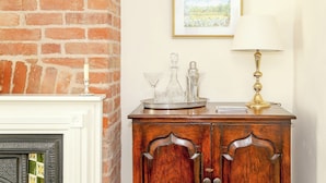 Une pièce avec un mur en briques, une cheminée blanche, un meuble en bois contenant de la verrerie, un tableau encadré et une lampe de table.