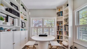 Una habitación con una mesa redonda, bancos, estanterías blancas y un amplio ventanal con persianas.