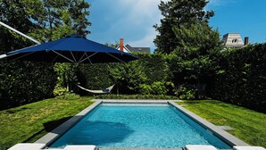 Una piscina rectangular con un suelo de color oscuro, rodeada de un césped cuidado y abundante vegetación. Hay sillas de descanso y una hamaca para relajarse.