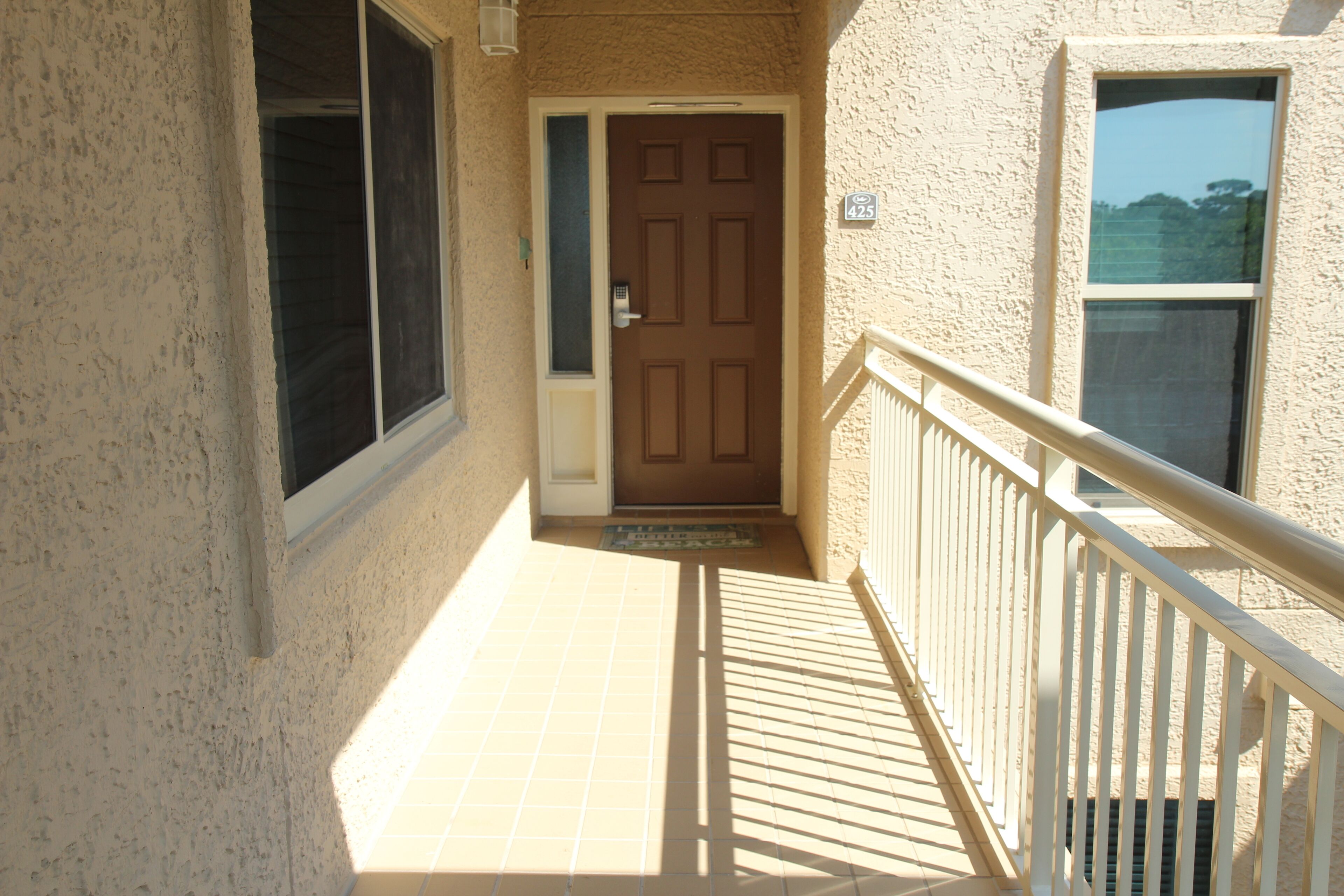 A narrow, sunlit corridor with a brown door, a window on the right, and a white railing.