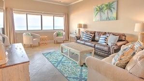 Ein Wohnzimmer mit einer Couch, Sesseln, einem Couchtisch und Blick auf das Meer durch große Fenster.