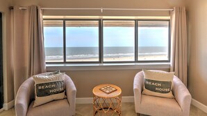 Ein Zimmer mit zwei Sesseln, die zu einem großen Fenster mit Blick auf den Strand hin ausgerichtet sind. Jedes Sitzmöbel hat ein Kissen mit der Aufschrift 'BEACH HOUSE'.