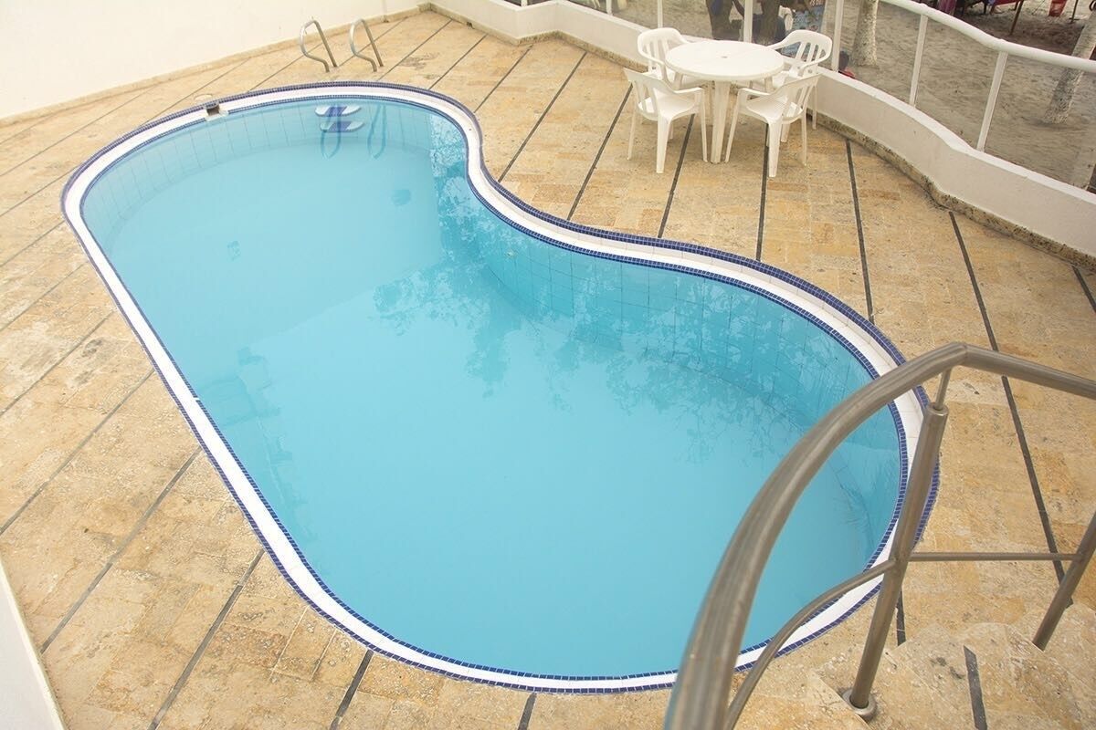 A swimming pool with a blue border, surrounded by a tiled patio area with white chairs and a small table.