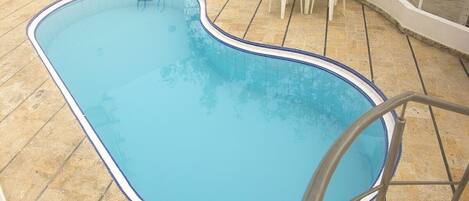 A swimming pool with a blue border, surrounded by a tiled patio area with white chairs and a small table.