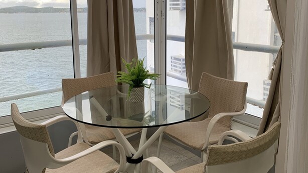 Ein Eckzimmer mit Glastisch und weißen Korbstühlen, einer Zimmerpflanze und Meerblick durch große Fenster.