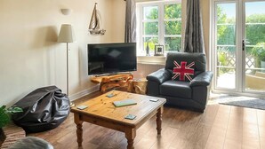 A living room with a flat-screen TV, a wooden coffee table, a black leather sofa, a black armchair with a Union Jack pillow, and a large window with a view of greenery. - 2 bedroom accommodation in Binfield, near Newport (Binfield, near Newport)