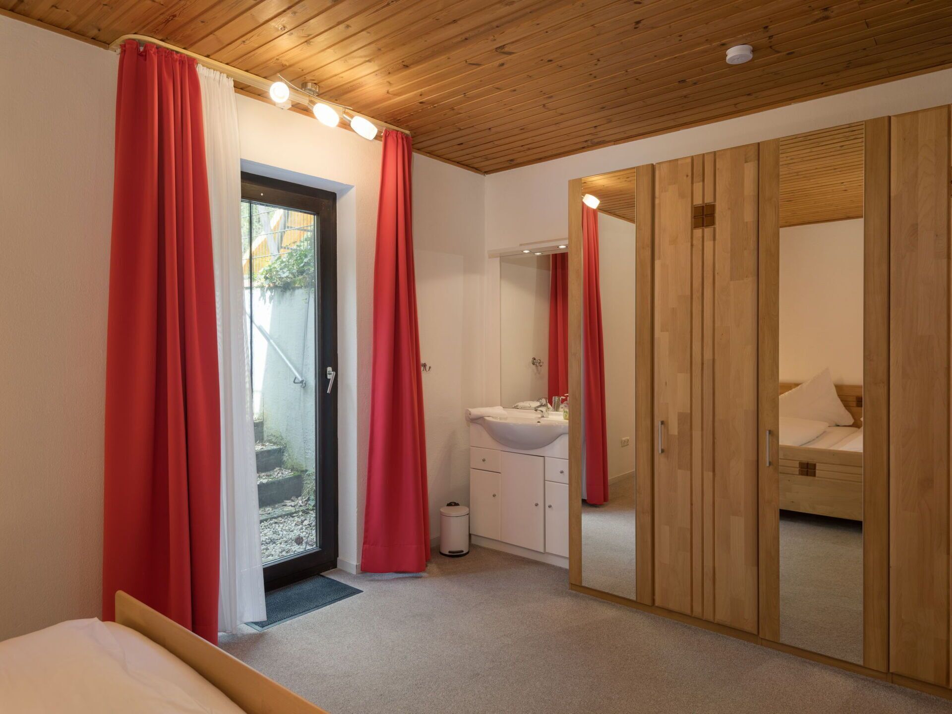 Une chambre avec un plafond en bois, un lit, une salle de bain avec un lavabo et un miroir, et des rideaux rouges.