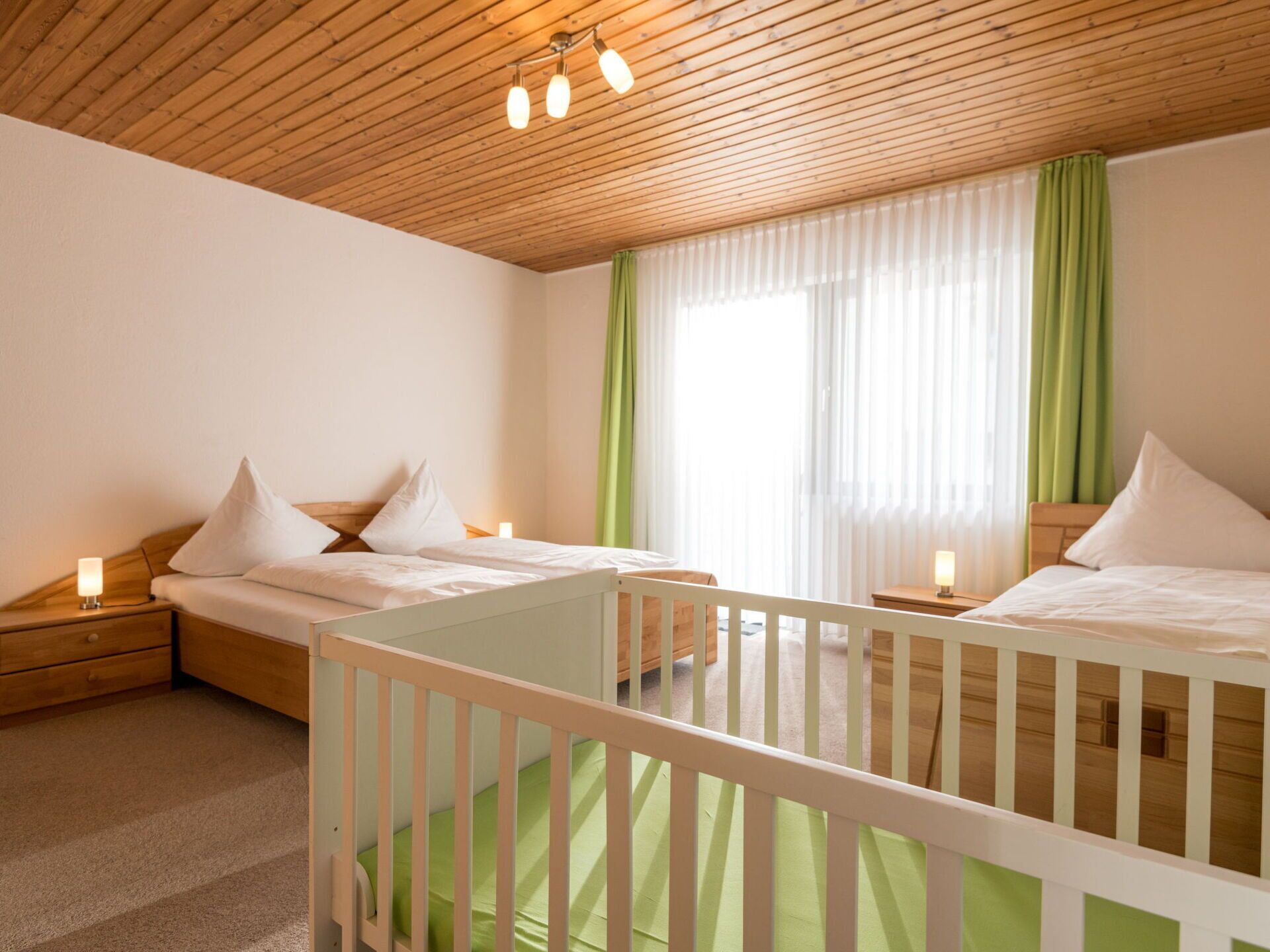 Een kamer met twee bedden, een ledikant, houten plafond en een raam met groene gordijnen.