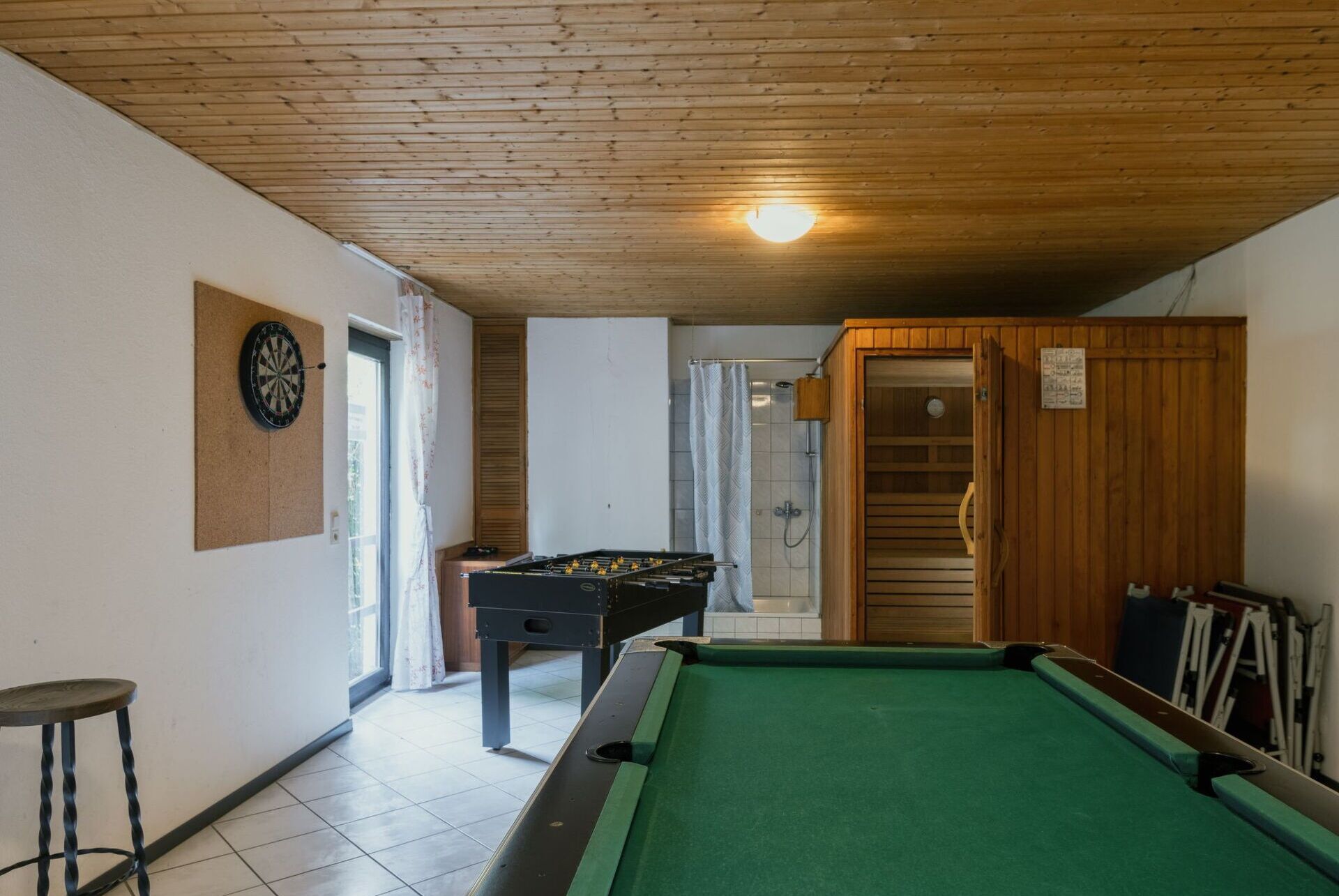 A room with a pool table, foosball table, and dartboard. There is a shower area with a wooden door and a towel rack.
