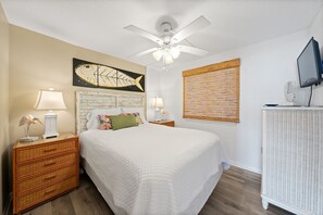 Une chambre à coucher comprenant un lit, deux tables de chevet, un ventilateur de plafond, un téléviseur fixé au mur et une tête de lit en bois.