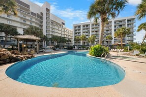 Une piscine entourée de chaises longues et de palmiers, avec un immeuble à plusieurs étages en arrière-plan.