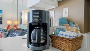 A coffee maker on a counter with a basket of snacks and a bag labeled "Young's Suncoast Realty." - Cozy Condo w/ Gulf Views | Boat-Friendly, Pool, Hot Tub, and Tennis Courts (Gulf Shores)