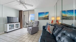 A living room with a television, a sofa, a chair, a ceiling fan, and artwork on the walls. - Cozy Condo w/ Gulf Views | Boat-Friendly, Pool, Hot Tub, and Tennis Courts (Gulf Shores)