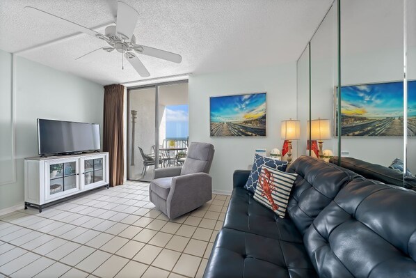 A living room with a television, a sofa, a chair, a ceiling fan, and artwork on the walls.