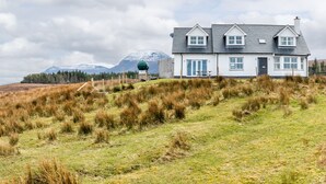 Una casa de dos pisos con fachada blanca y techo oscuro, ubicada en una colina cubierta de hierba con una montaña al fondo.