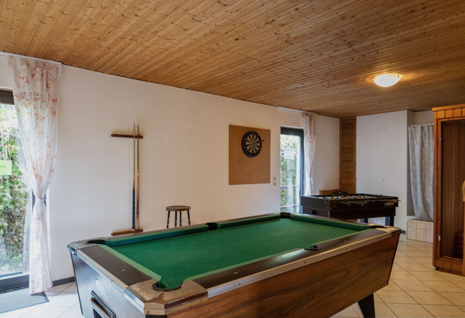 Mesa de bilhar numa sala com teto de madeira, um alvo de dardos e uma mesa de futebol de mesa.