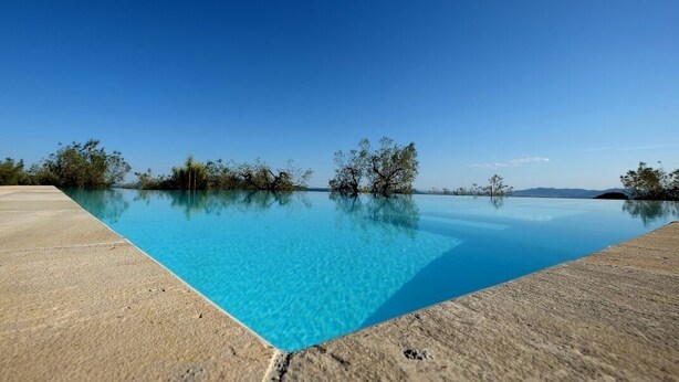 A clear blue infinity pool with a concrete edge, surrounded by sparse trees and a clear sky. - Agriturismo Campo al Crognolo. (Sassofortino)