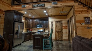 A wooden cabin interior with a kitchen featuring dark cabinets, a refrigerator, a microwave, and a sink. A welcome sign hangs above the kitchen, and a sign on the wall reads "Life Better at the Cabin."