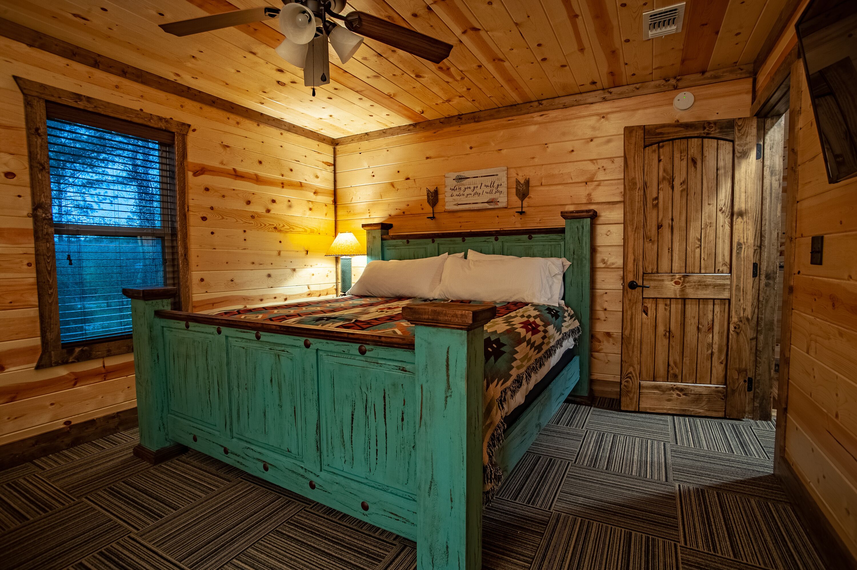 A wooden cabin bedroom with a bed, a ceiling fan, a window with blinds, and a door.