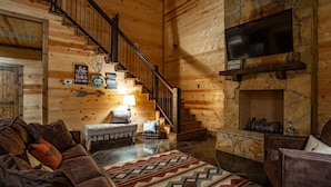 A cozy log cabin interior with a wooden staircase, a fireplace, and a leather sofa.