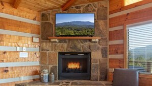 Un acogedor interior de cabaña con chimenea de piedra, un televisor montado encima y un sillón cómodo junto a una ventana.