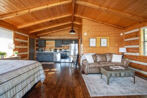 Un interior acogedor de cabaña con techo de madera, una kitchenette, una sala de estar con sofá y mesa de centro, y una cama con colcha a rayas.