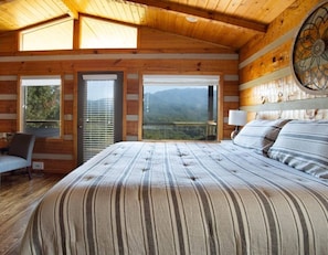 Quarto com uma cama grande, uma cadeira, uma escrivaninha e vista para montanhas pela janela.