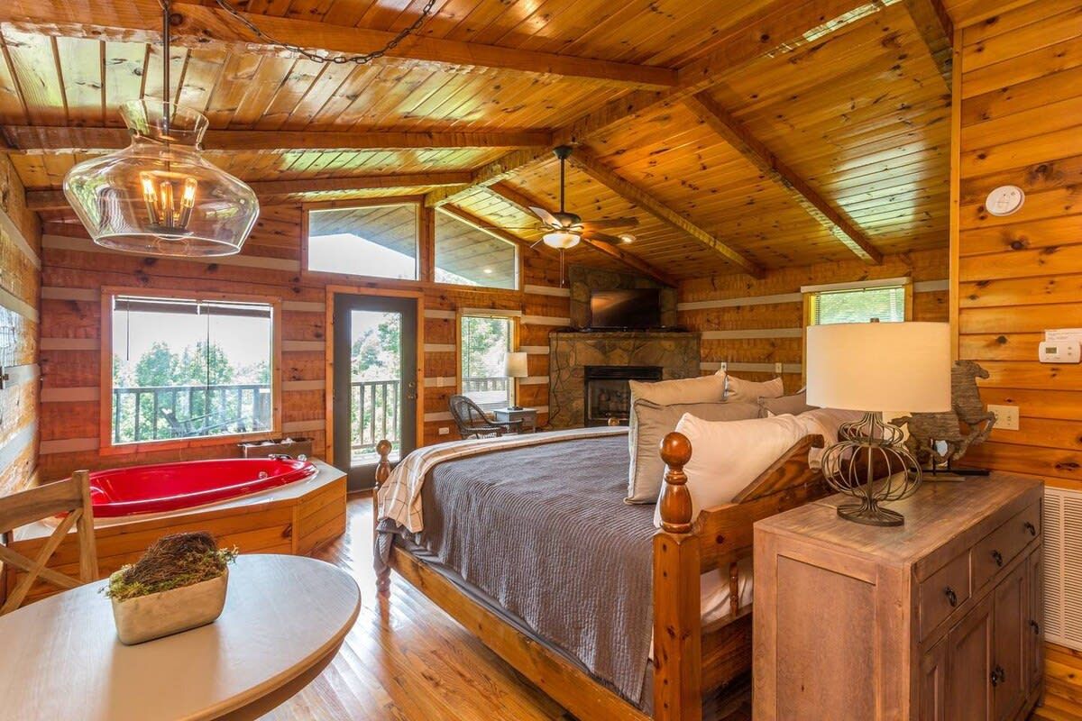 A log cabin bedroom with a large bed, a red bathtub, a fireplace, and a balcony.