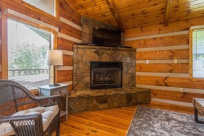 Interior de una cabaña de troncos con chimenea de piedra, paredes de madera, un sillón de mimbre y un televisor sobre la chimenea.