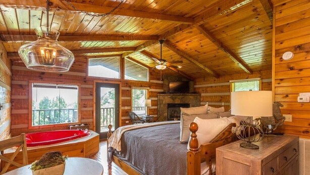 Ein Schlafzimmer in einer Blockhütte mit einem großen Bett, einer roten Badewanne, einem Kamin und einem Balkon mit Blick auf Bäume.
