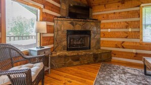 Ein gemütliches Blockhaus-Interieur mit Kamin aus Stein, Holzwänden, einem Korbsessel und einem kleinen Tisch.