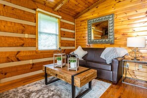 Interior acogedor de una cabaña con pared de madera, una ventana con persianas, un sofá, una mesa de centro con faroles y un espejo.
