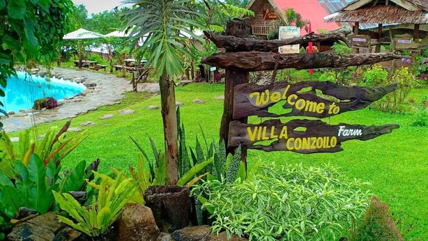 Un senyal de fusta amb la inscripció 'Welcome to Villa Conzoi' i 'Farm', envoltat de vegetació exuberant i una piscina.