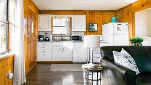 Une cuisine compacte avec des meubles blancs, un four à micro-ondes et un réfrigérateur. Il y a une fenêtre avec des rideaux, un petit tabouret et un canapé en cuir noir avec un coussin.