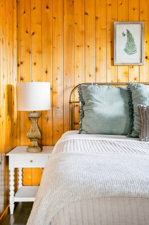 Un dormitorio con pared de madera, una cama con colcha blanca, una mesita de noche con lámpara y una obra de arte con helecho enmarcado.