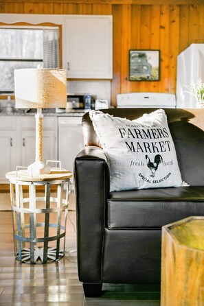 Una sala de estar acogedora con un sofá de cuero negro, una mesita redonda de metal con una lámpara y un cojín con el texto 'Farmers Market'.