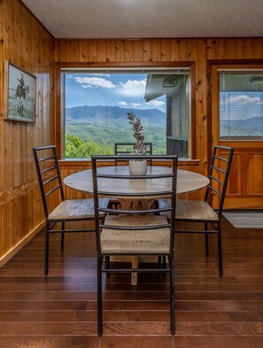 A wooden-paneled room with a round table and chairs, a window with a mountain view, and a framed picture on the wall.