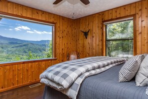 Un dormitorio con un ventanal grande, paredes de madera, una cama con ropa de cuadros y un ventilador de techo.