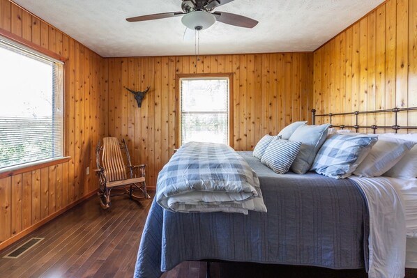 Una habitación con paredes de madera, una cama con cubrecama de cuadros, un ventilador de techo y una ventana con persianas.