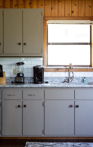 A kitchen with grey cabinets, a window, a sink, a blender, and a coffee maker.