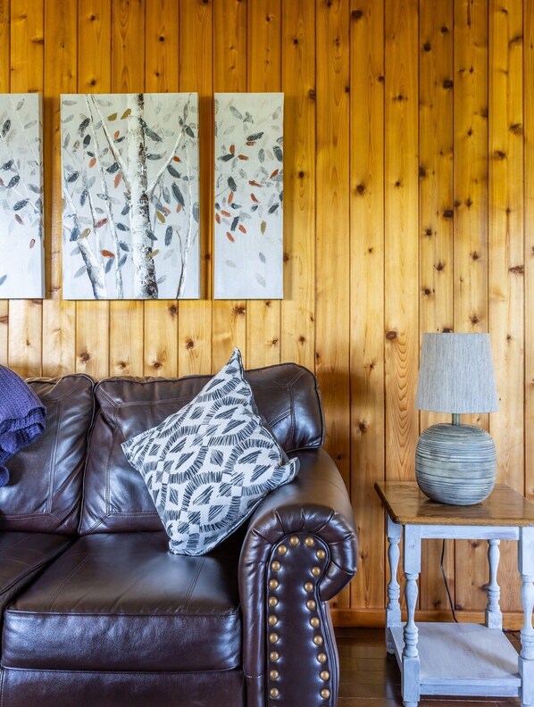 A brown leather sofa with patterned cushions, a wooden wall with three abstract paintings, and a small table with a lamp.