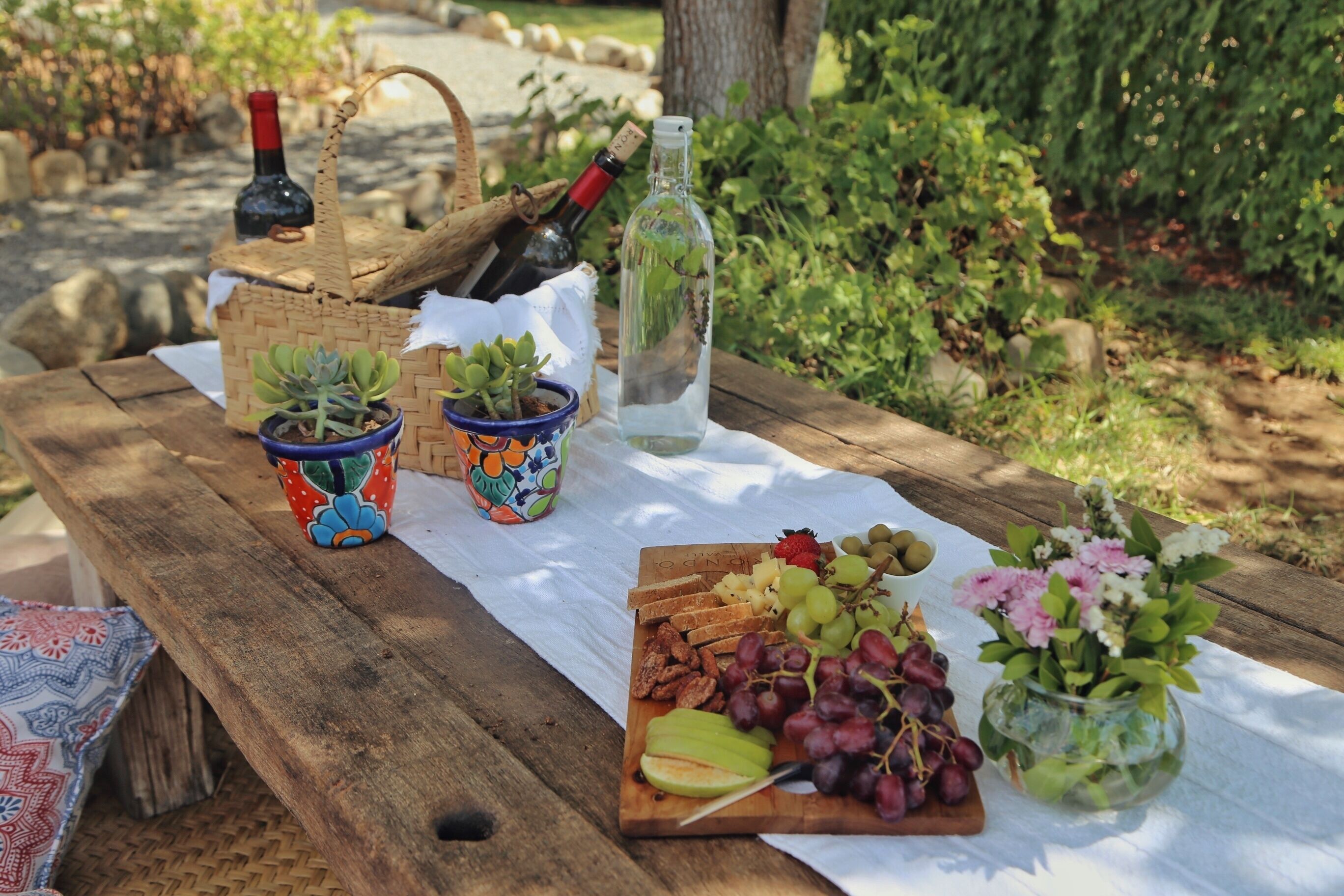 Un montaje para picnic con una mesa de madera, diversas frutas, uvas, una botella de vino y pequeñas macetas.